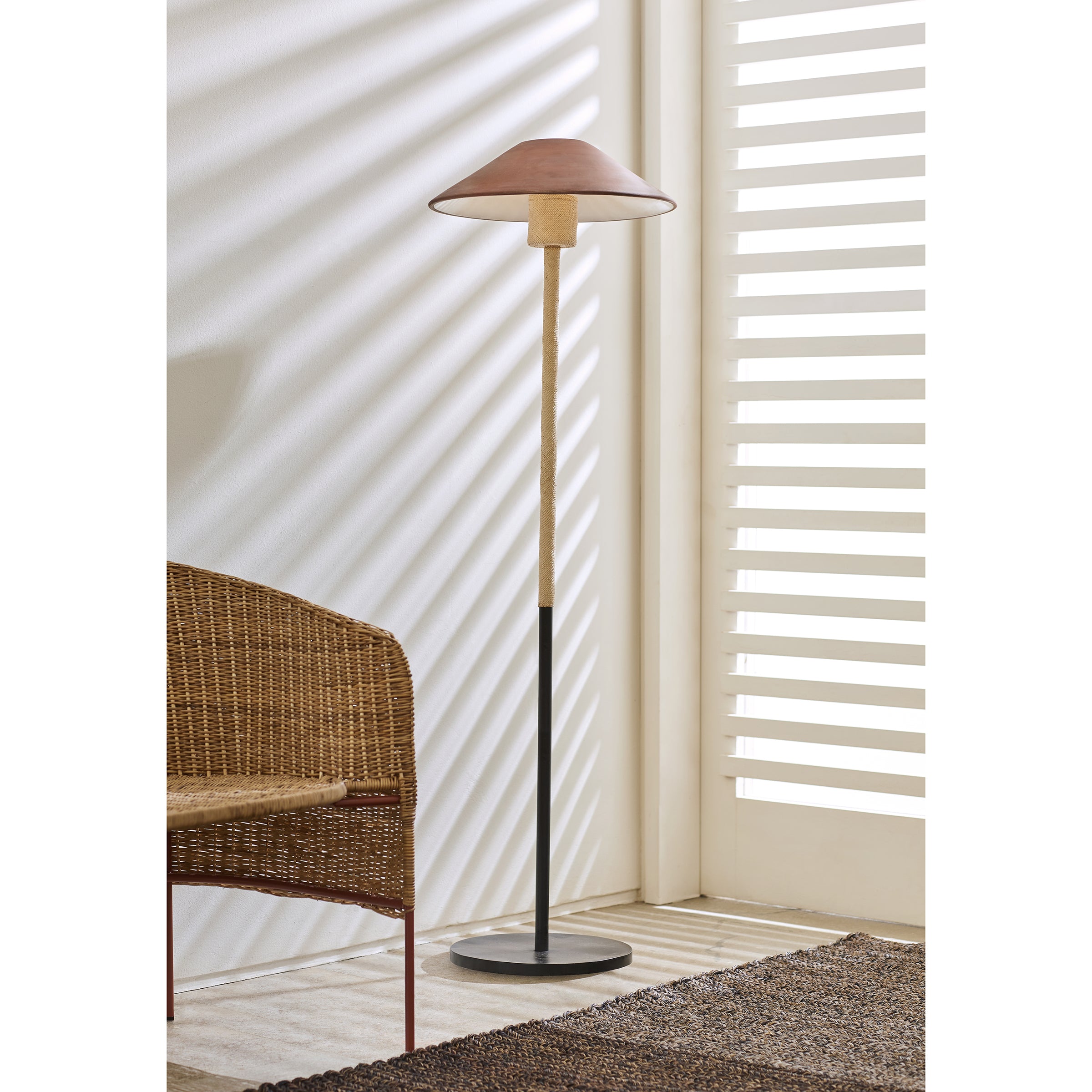 The Ames Raiz Floor Lamp by Ames, featuring a brown shade and wooden stand, stands beside a wicker chair. Sunlight filters through white blinds, casting shadows and showcasing the beauty of Colombian craftsmanship throughout.