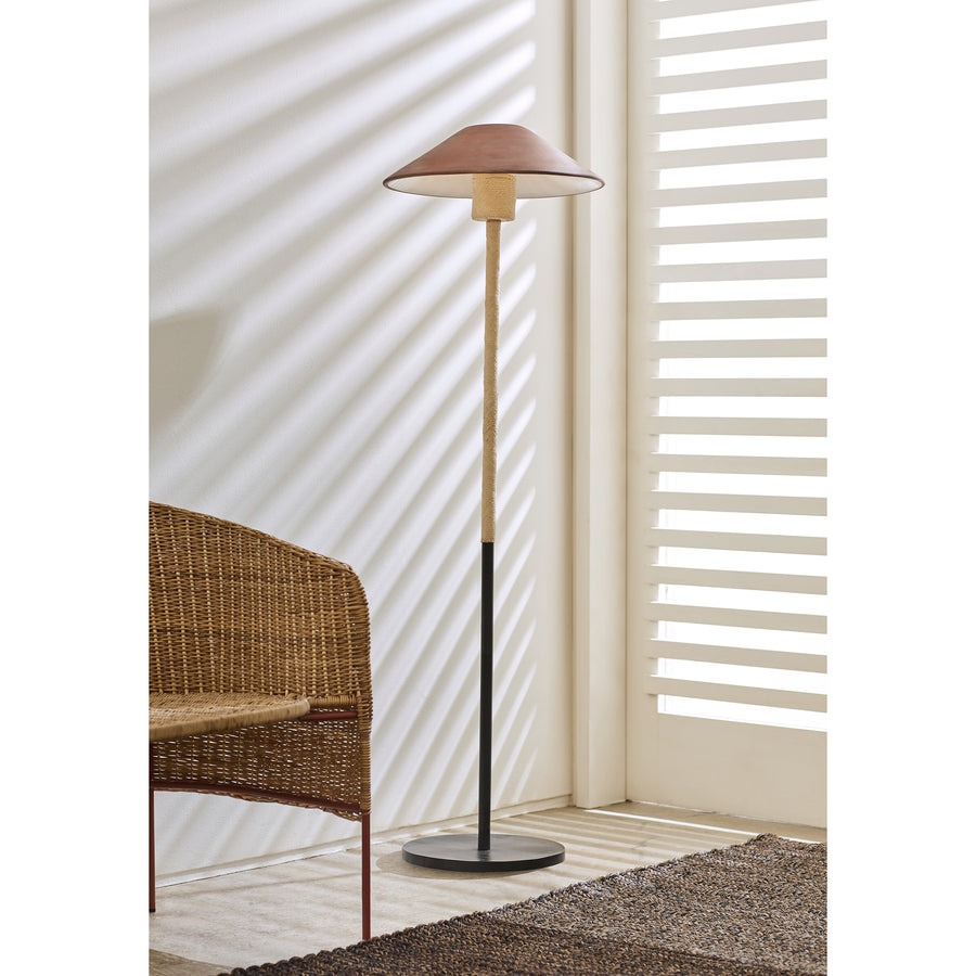 The Ames Raiz Floor Lamp by Ames, featuring a brown shade and wooden stand, stands beside a wicker chair. Sunlight filters through white blinds, casting shadows and showcasing the beauty of Colombian craftsmanship throughout.