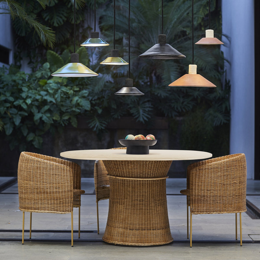 A round wicker table with four wicker chairs sits outdoors beneath several Ames Raiz Pendant Lamp Smalls by Ames, each handmade to showcase Colombian craftsmanship. Lush green plants and trees in the background create a cozy, natural setting.