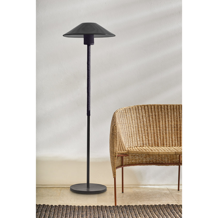 The Ames Raiz Floor Lamp by Ames, featuring a wide flat shade and modern black design, stands next to a wicker chair against a plain white wall, showcasing minimalist style and Colombian craftsmanship.