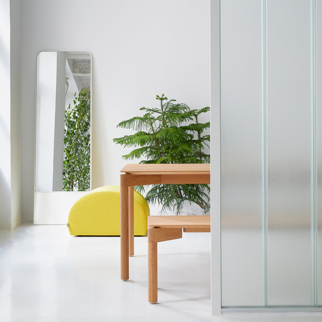 A modern room showcases an Objekte unserer Tage Wedekind Bench made from sustainably certified oak next to a frosted glass divider. A tall mirror reflects greenery, while a yellow cushioned seat provides a splash of color. Bright natural light emphasizes the precision craftsmanship and minimalist decor.