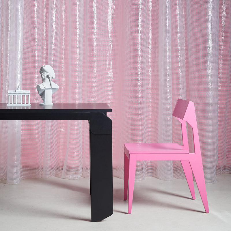 A modern black table and a durable Schulz Chair in pink by Objekte unserer Tage, crafted from solid ash wood, are set on a light floor with sheer pink curtains. A white bust and miniature architectural model adorn the table, creating a minimalist, artistic setting that reflects SCHULZ design sophistication.