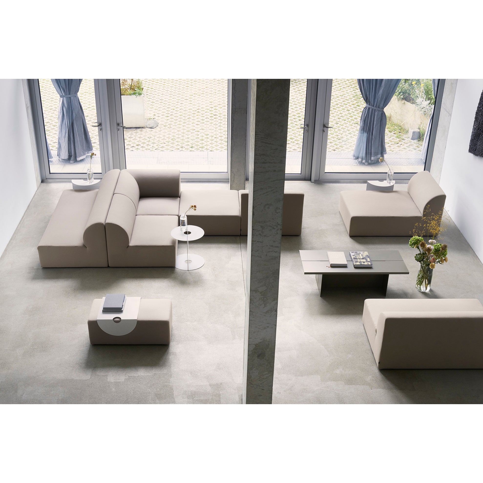 A modern living room features Objekte unserer Tages Weber Modular Sofas around a rectangular coffee table. A round side table with geometric decor stands nearby, while large windows with blue curtains offer outdoor views. A bouquet of flowers graces the console table.