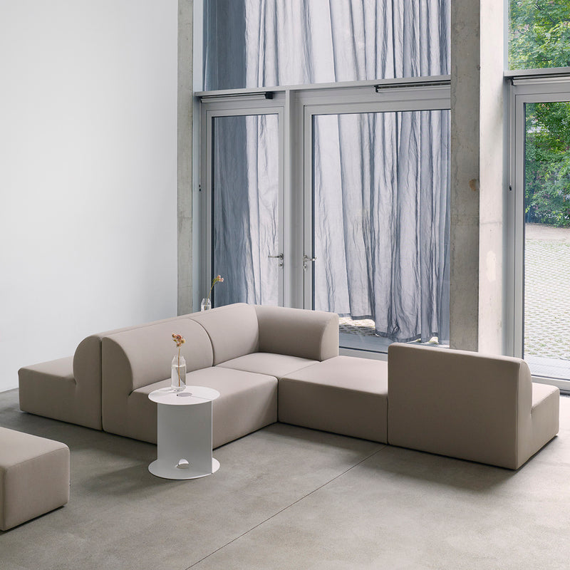 The Weber Modular Sofa by Objekte unserer Tage, featuring a minimalist beige design and geometric shapes, forms an L-shape. In front is a small round white table with a flower vase. Large windows with sheer curtains provide views of the greenery outside.