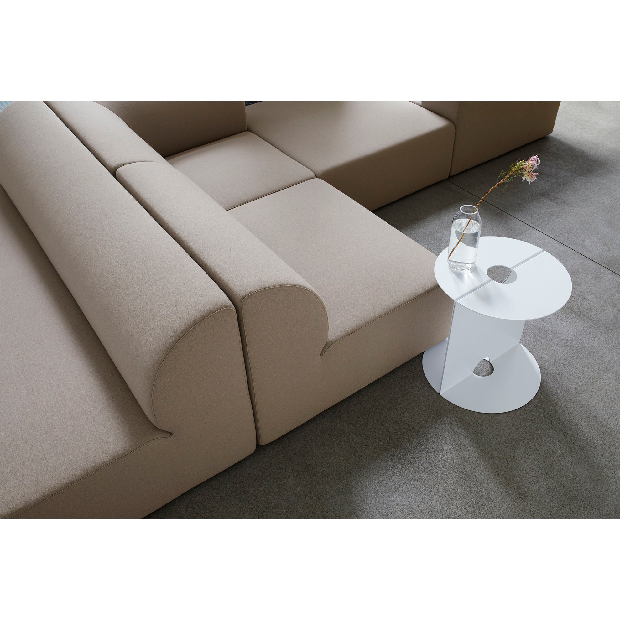 A minimalist living room showcases an Objekte unserer Tage Weber Modular Sofa and a sleek white round side table adorned with a glass vase holding a pink flower. A neutral-toned rug completes the space, adding German sensuality.