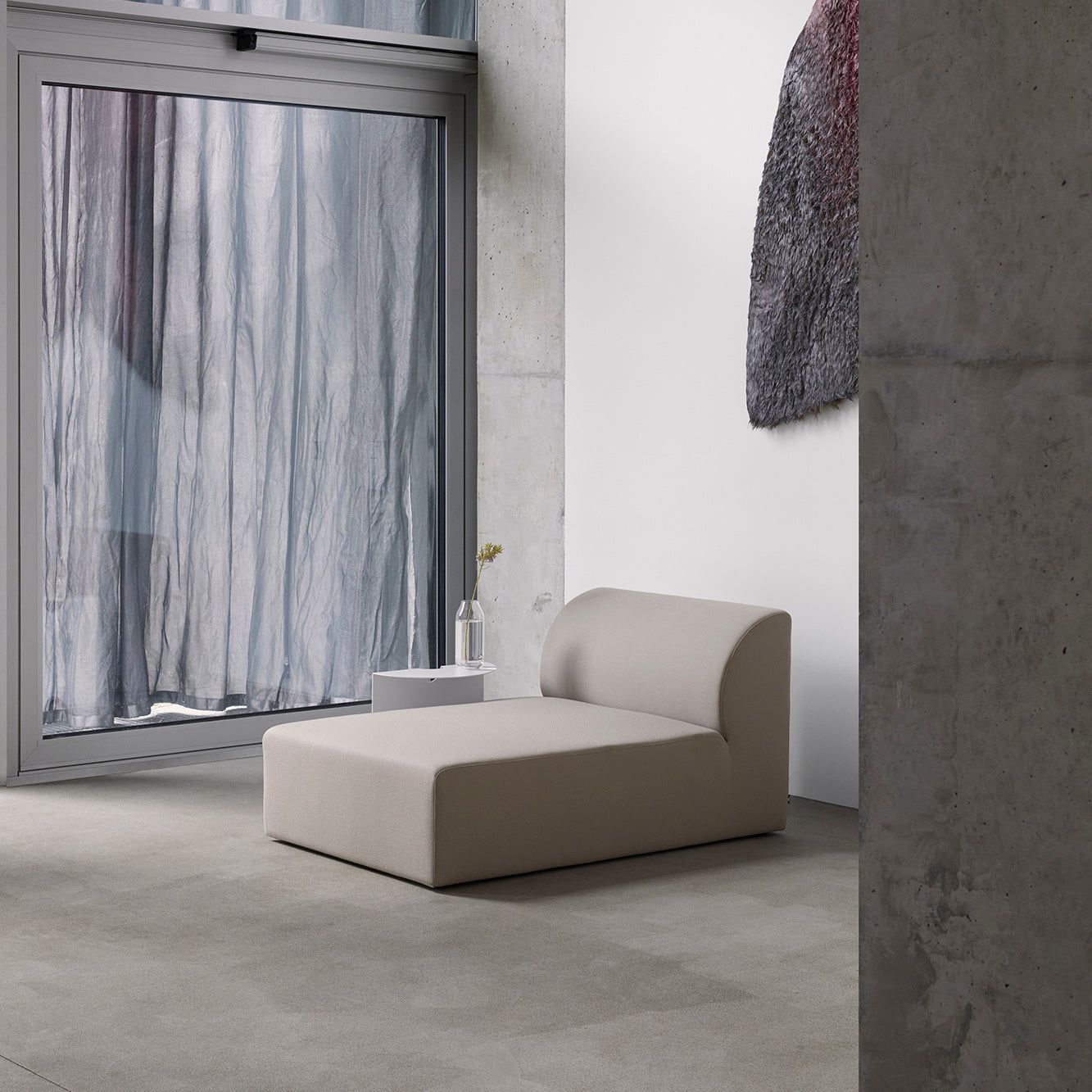 The minimalist room features a beige Weber Modular Sofa by Objekte unserer Tage and a small side table holding a vase with a single flower. Nearby, textured wall art adds intrigue while large windows with sheer curtains filter soft light, enhancing the spaces German sensuality.