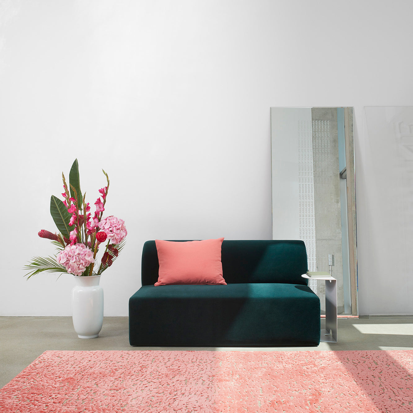 A modern living room features an Objekte unserer Tage Weber Modular Sofa in dark green with a pink cushion. A large mirror leans against the white wall, reflecting sustainable awareness. Pink and red flowers in a vase adorn the left, while a patterned pink rug adds German sensuality to the floor.