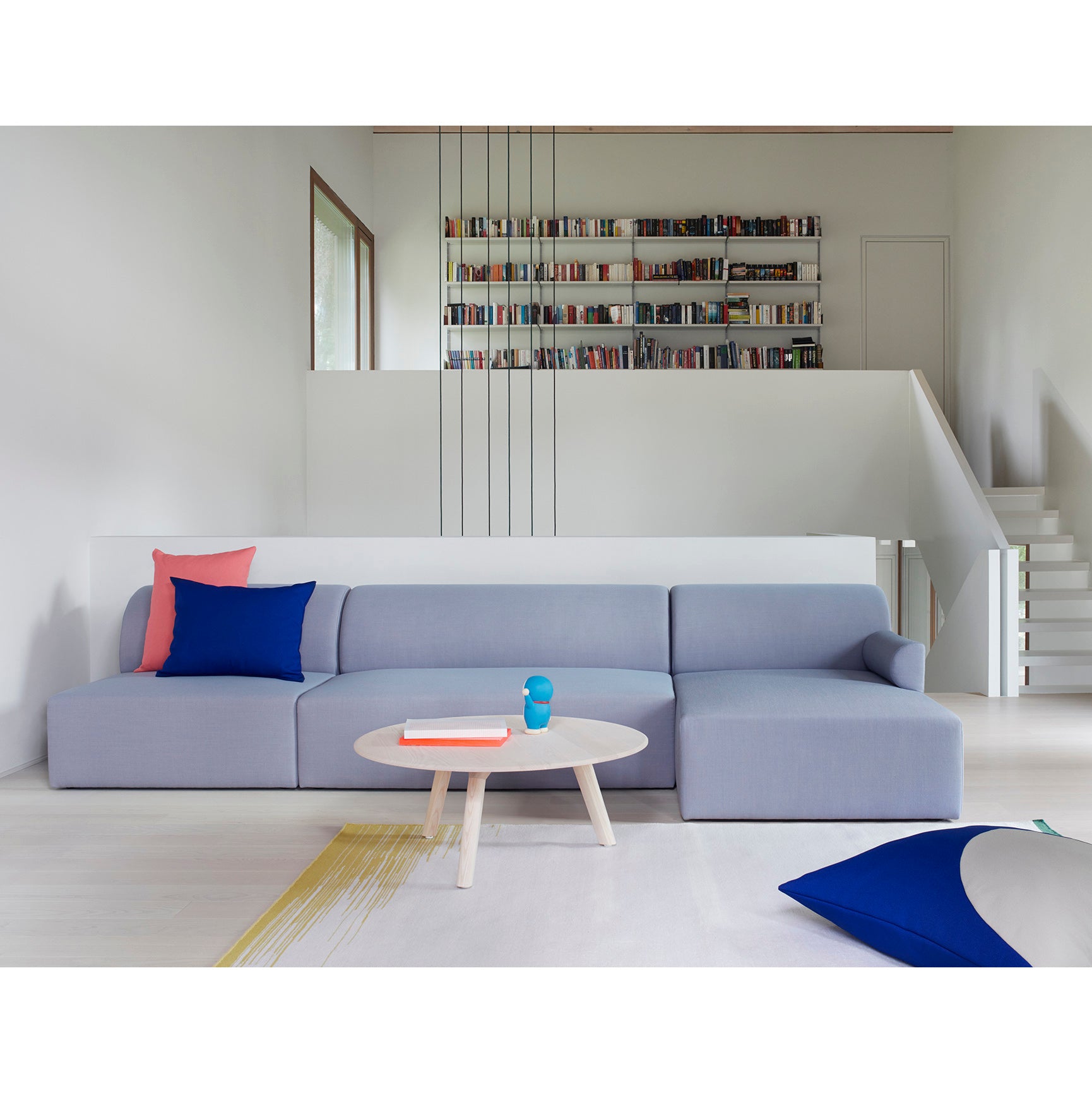 A modern living room showcases a light gray Weber Modular Sofa by Objekte unserer Tage, enhanced with a pink and blue cushion. In front, a round wooden coffee table holds books and a blue cup. A wall-mounted bookshelf and white staircase in the background boost sustainable awareness.