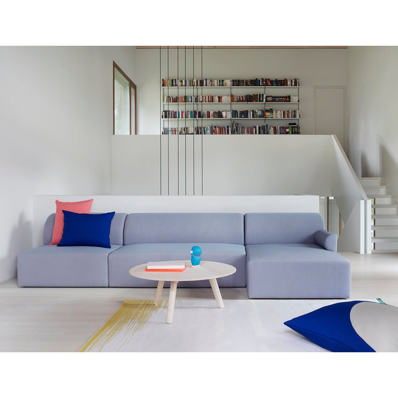 A modern living room showcases a light gray Weber Modular Sofa by Objekte unserer Tage, enhanced with a pink and blue cushion. In front, a round wooden coffee table holds books and a blue cup. A wall-mounted bookshelf and white staircase in the background boost sustainable awareness.