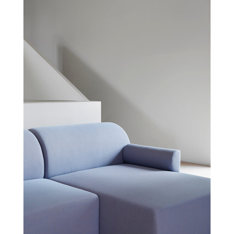 The minimalist room showcases an Objekte unserer Tage Weber Modular Sofa in blue, featuring rounded edges and a cylindrical armrest cushion. A light gray wall, complemented by soft natural light, creates a serene atmosphere that embodies German sensuality and sustainable awareness with its elegant design.