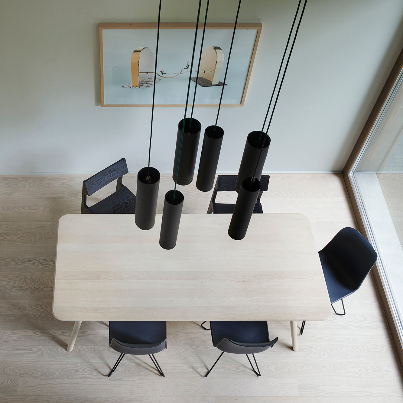 The dining area showcases the Meyer Table Round by Objekte unserer Tage in solid ash, paired with six black chairs and black cylindrical pendant lights above. A framed abstract artwork adorns the light-colored wall, enhancing its modern elegance.