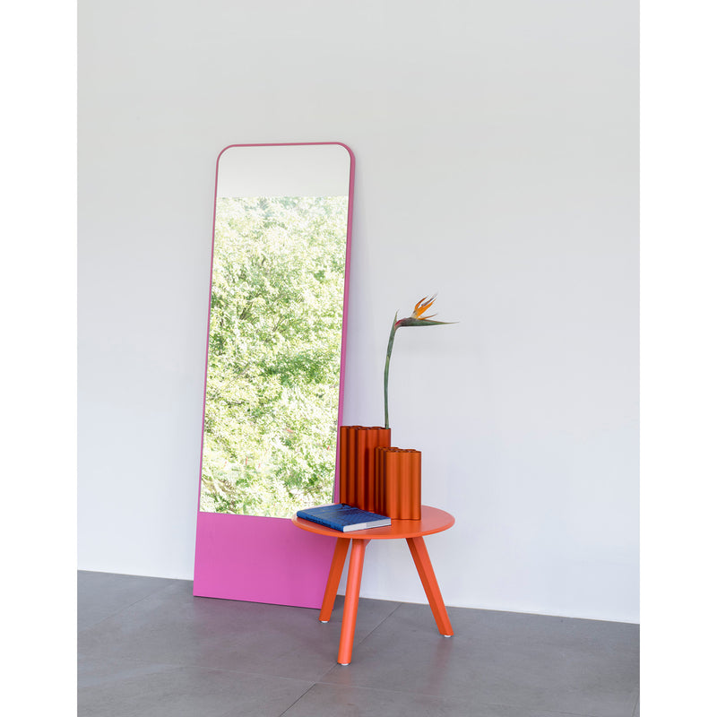 The Friedrich Mirror by Objekte unserer Tage, with its tall pink frame, reflects lush green foliage, enhancing the minimalist design of this contemporary living space. It leans against a white wall beside an orange table holding blue books and a vase with a bird of paradise flower on the smooth gray floor.