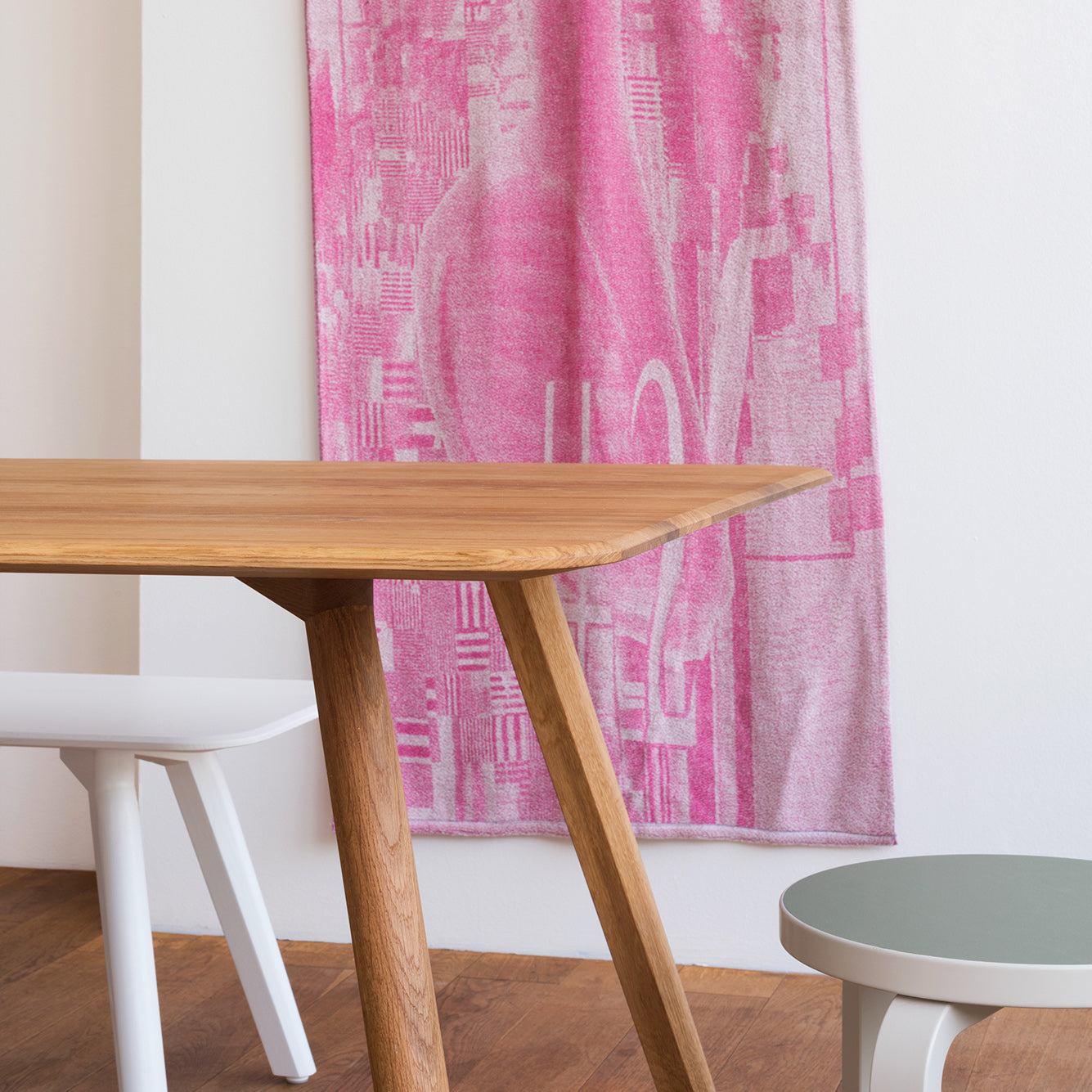 The MEYER Table Round by Objekte unserer Tage, crafted from solid ash wood with angled legs, stands gracefully on a wooden floor. A white stool peeks beside it, while a pink patterned textile adorns the wall. On the right, an elegantly designed small round table with a green top completes the scene.