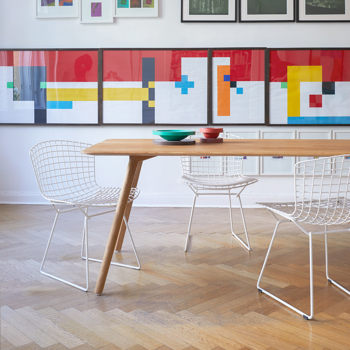 A modern dining room showcases an Objekte unserer Tage Meyer Table Round crafted from solid ash wood, paired with white wire chairs on a herringbone floor, with vibrant abstract artwork featuring red, blue, and yellow geometric shapes adorning the wall behind.