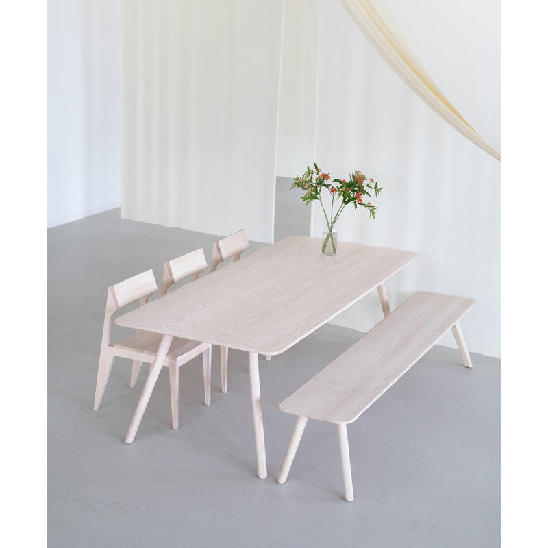 A minimalist dining area features a light wood table made of solid ash paired with the Meyer Bench and two matching chairs from Objekte unserer Tage. A vase of flowers atop the table enhances the serene atmosphere against a soft white backdrop.