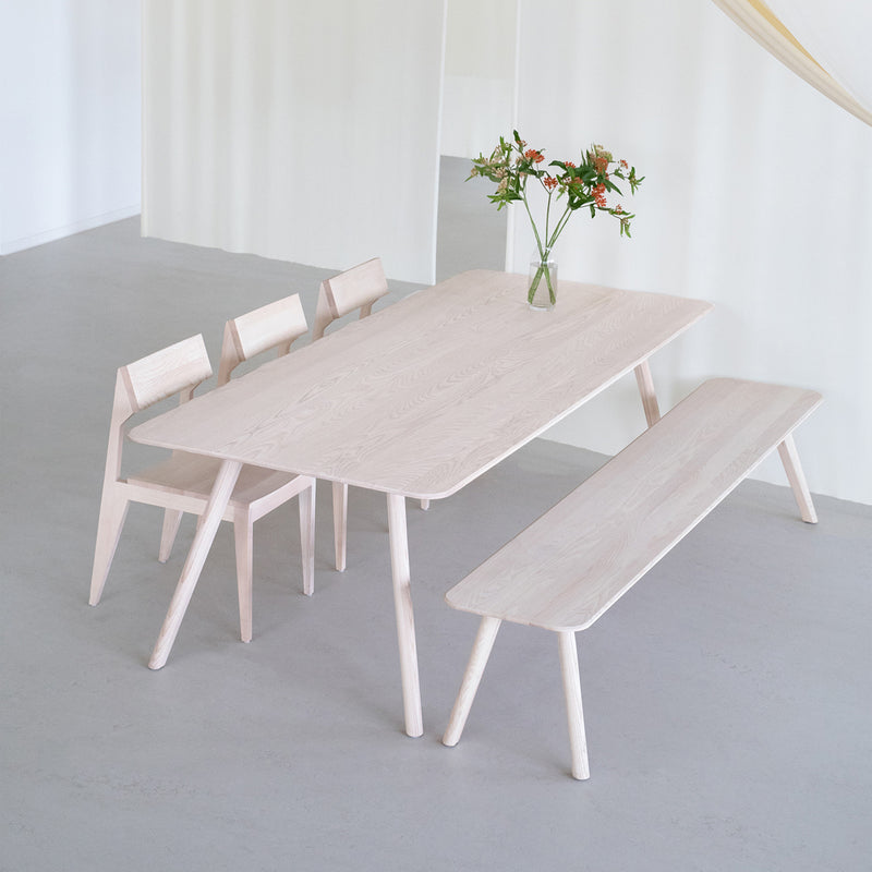 The minimalist dining area features an Objekte unserer Tage Meyer Table crafted from solid ash, complemented by three matching chairs and a bench. A glass vase with flowers adorns the table, enhancing the modern look with its clean lines and neutral color palette.