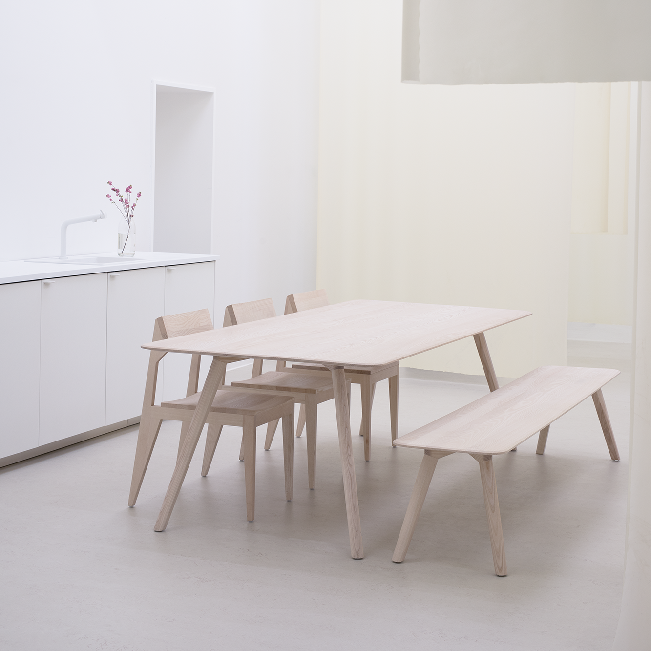 A cozy dining area with a solid ash table, two chairs, and the Objekte unserer Tage Meyer Bench. A white counter with a sink completes the look, enhanced by a small vase of red flowers. The room is illuminated in soft white and cream tones.