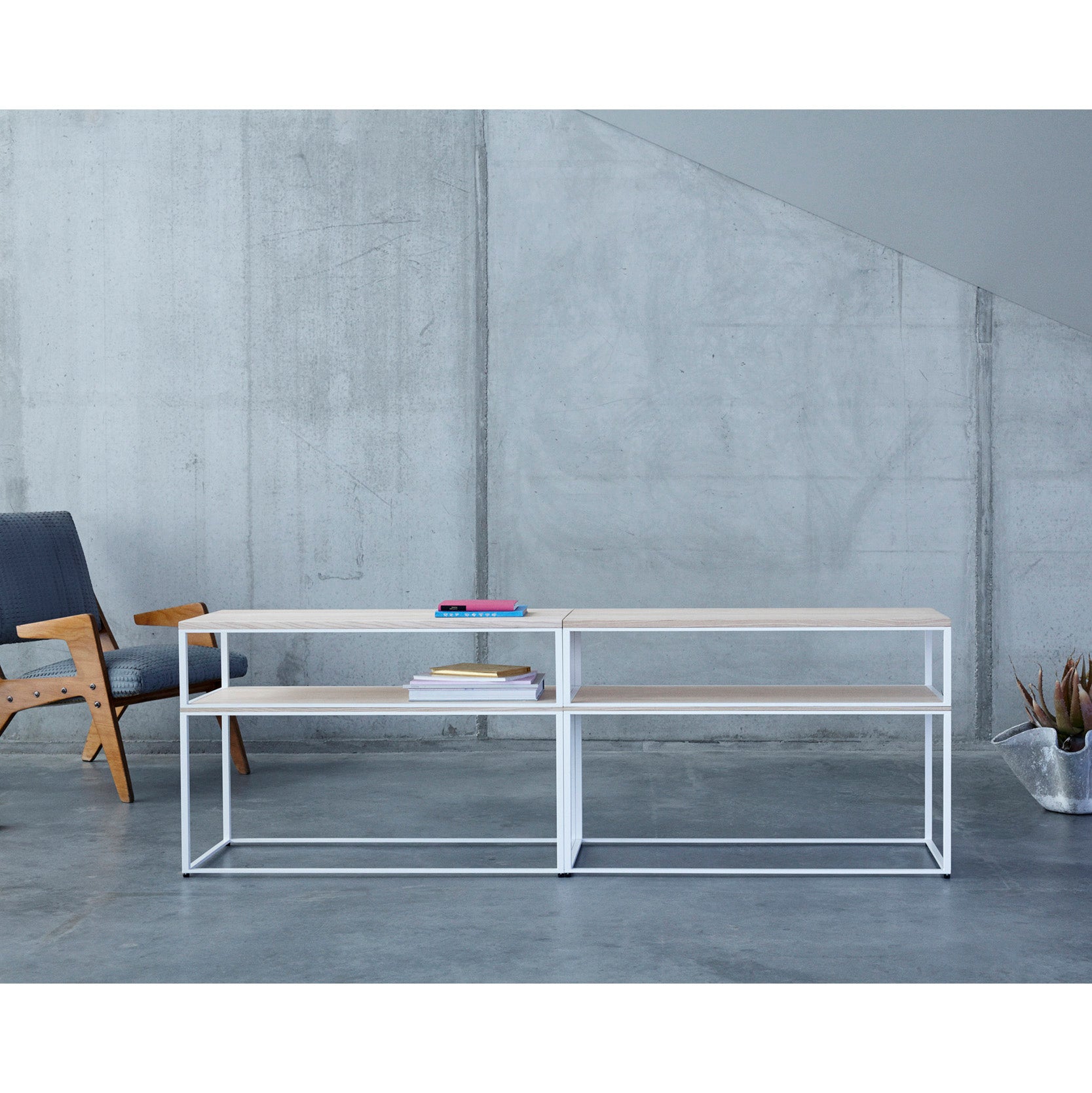 The minimalist room showcases a concrete wall with a wooden chair and blue cushion on the left. At the center is an Objekte unserer Tage Fischer Shelving System, featuring a white frame and wood top, adorned with several books. A potted plant rests on the floor to the right.