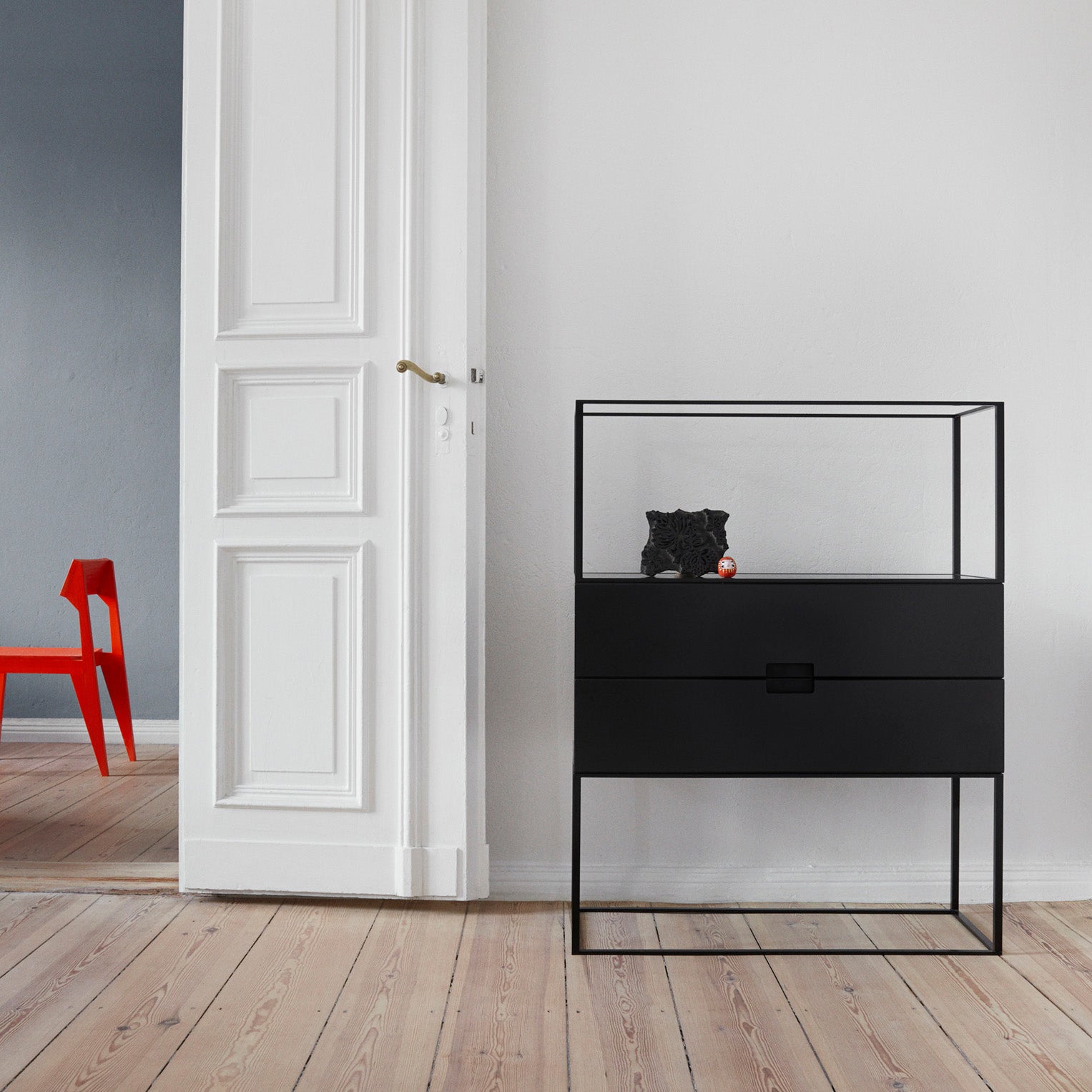 In a modern, minimalist room with light wood floors, the Fischer Shelving System by Objekte unserer Tage features modular furniture. A black cat sits inside its sleek black cabinet with glass sides. A red chair contrasts against a gray wall near tall white double doors, embodying puristic design.