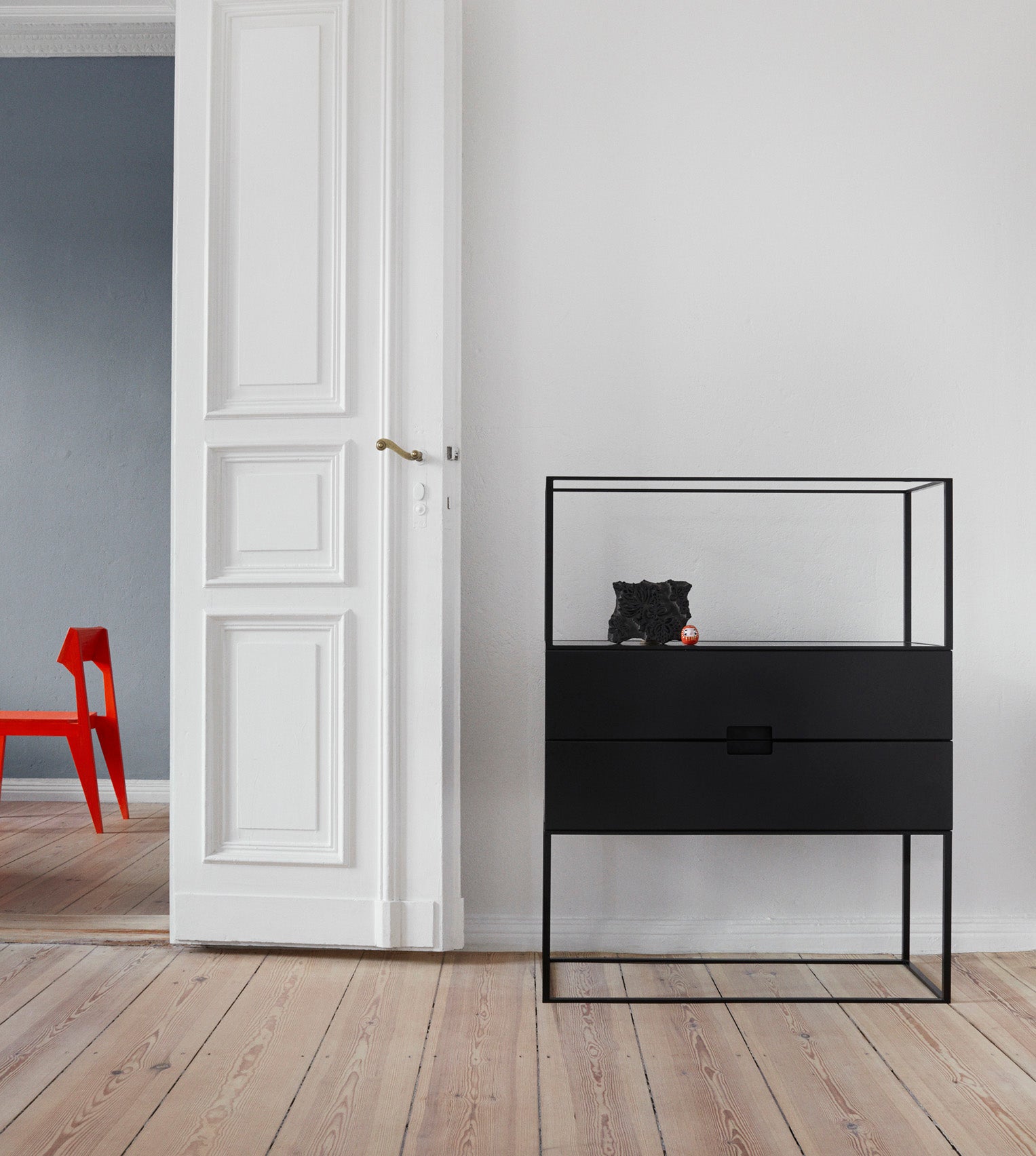 A minimalist room showcases a sleek Fischer Shelfing system by Objekte unserer Tage. A white door opens to a blue wall, with a red chair partially visible. The black cabinet sits on a light wooden floor, displaying decorative black and small red items.