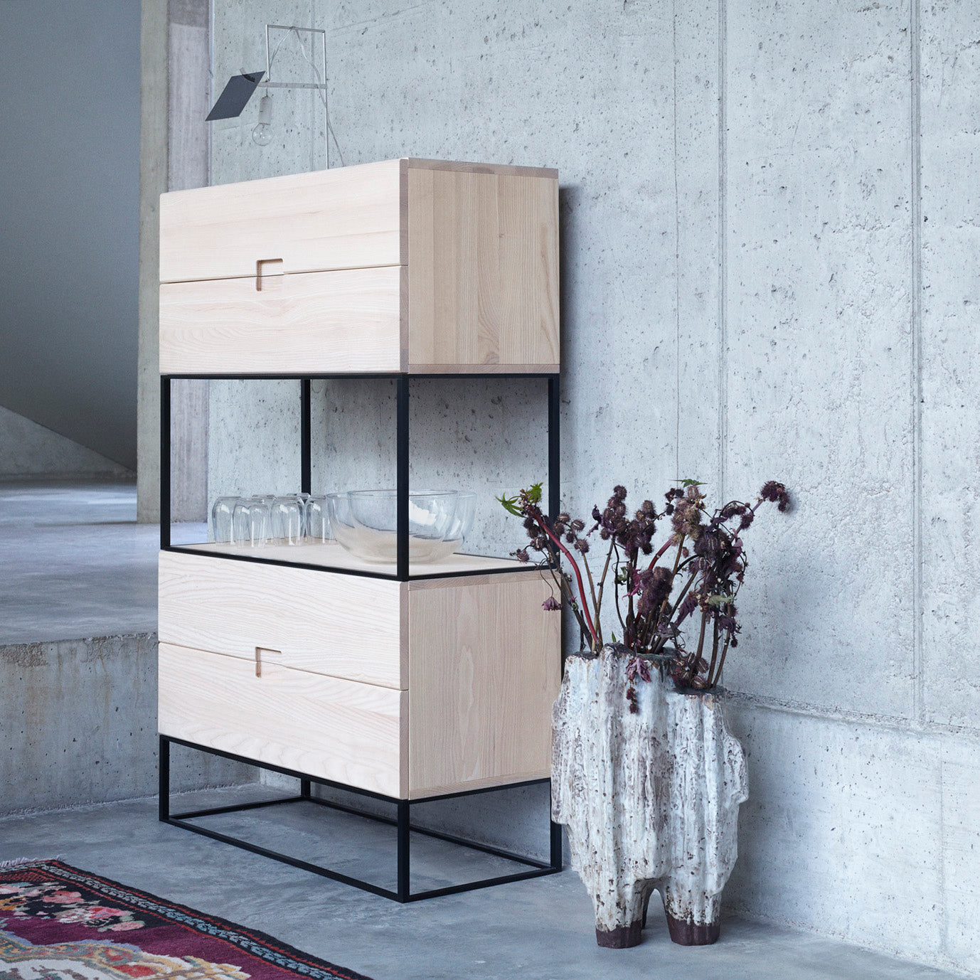 The Fischer Shelving System by Objekte unserer Tage, featuring a modern wooden design with a black metal frame, stands against a concrete wall. Below, glassware is on an open shelf alongside a textured vase with dried flowers on a colorful rug, perfectly complementing the industrial space.