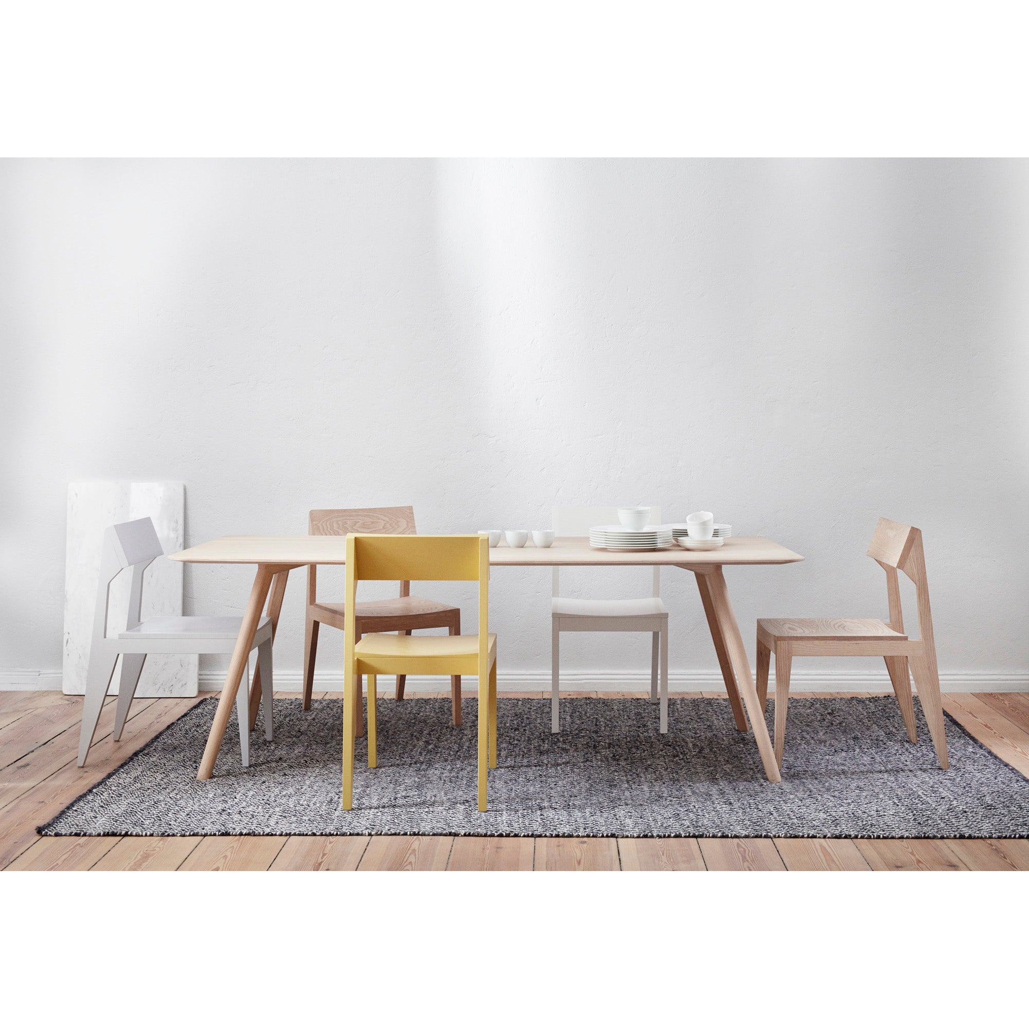 A minimalist dining room showcases an elegant design with the Meyer Table Round by Objekte unserer Tage, paired with assorted chairs, including a yellow one. It features white tableware on a grey textured rug on a wooden floor against a plain white wall.
