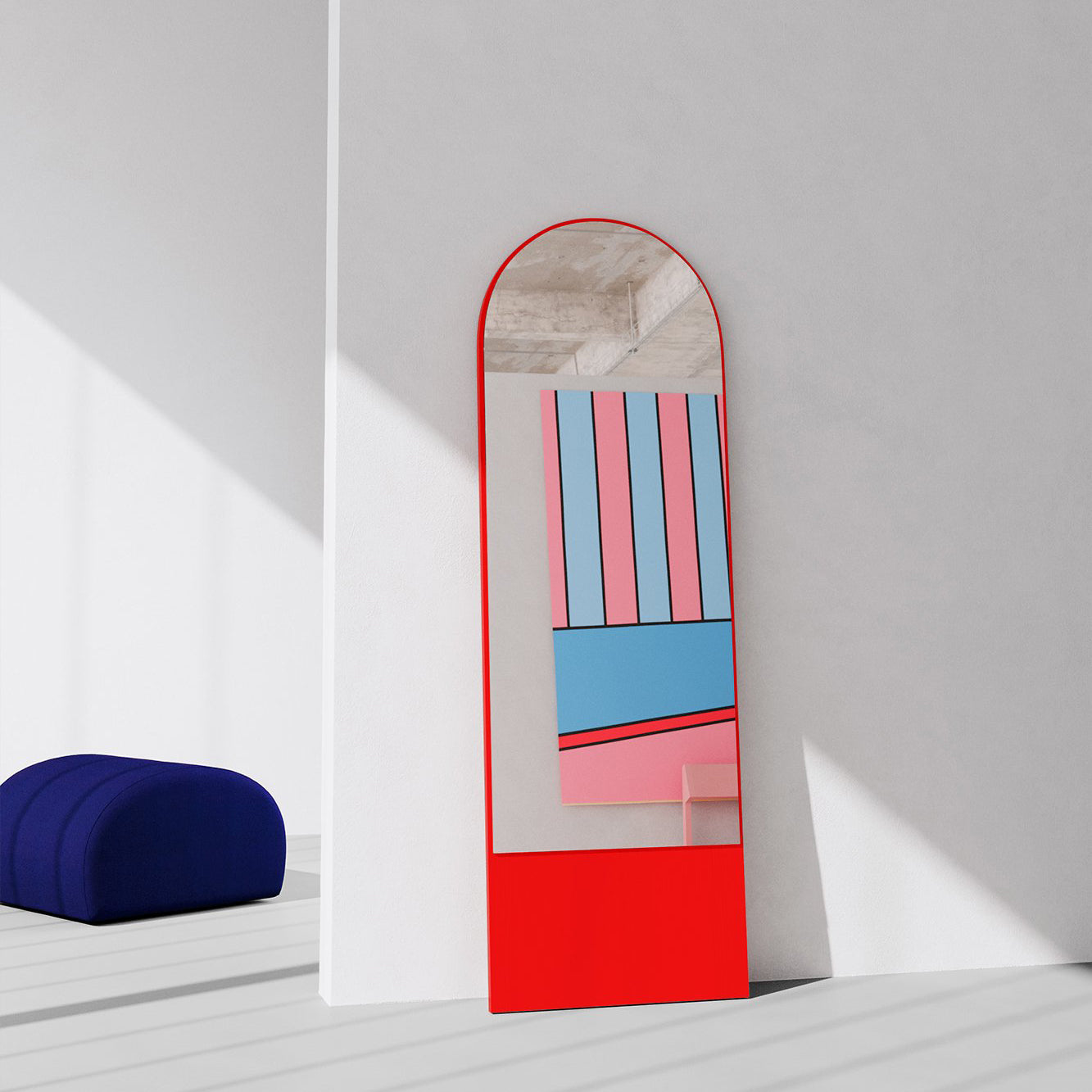 A tall, red-framed Objekte unserer Tage FRIEDRICH 21 Mirror with a round arch top leans against a white wall, reflecting colorful abstract patterns in pink, blue, and black. A dark blue rounded ottoman is on the left in the sunlit minimalist room.