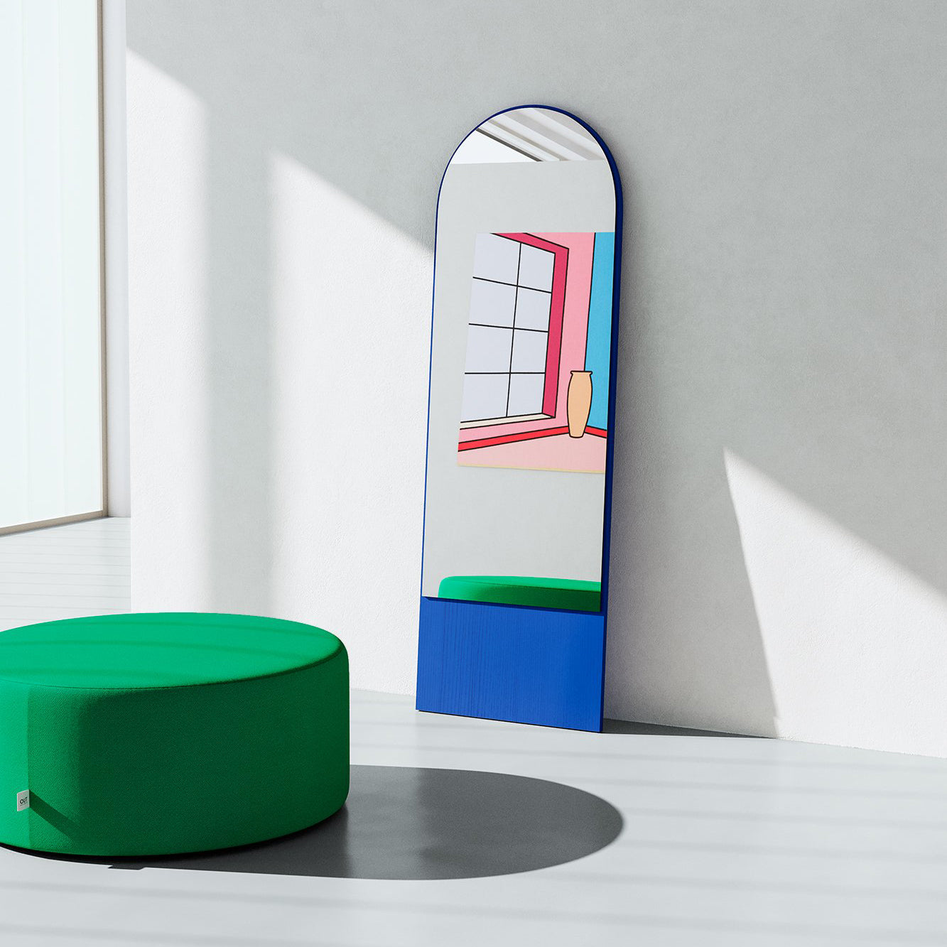 The FRIEDRICH 21 Mirror by Objekte unserer Tage, featuring colorful geometric patterns and a tall rectangular shape, reflects a window and vase. It leans against the wall in a minimalist room with a green circular ottoman, sunlight warming the lacquered ash wood accents.