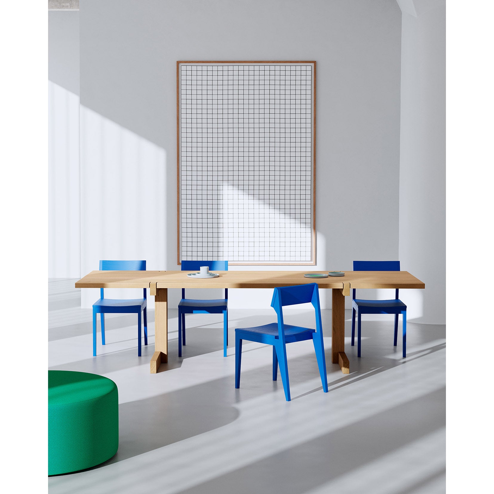 A modern room showcases an Objekte unserer Tage Richter Table made from sustainable forestry, surrounded by blue and white chairs. Sunlight highlights a large framed grid-patterned artwork on the wall while a green ottoman sits in the foreground.