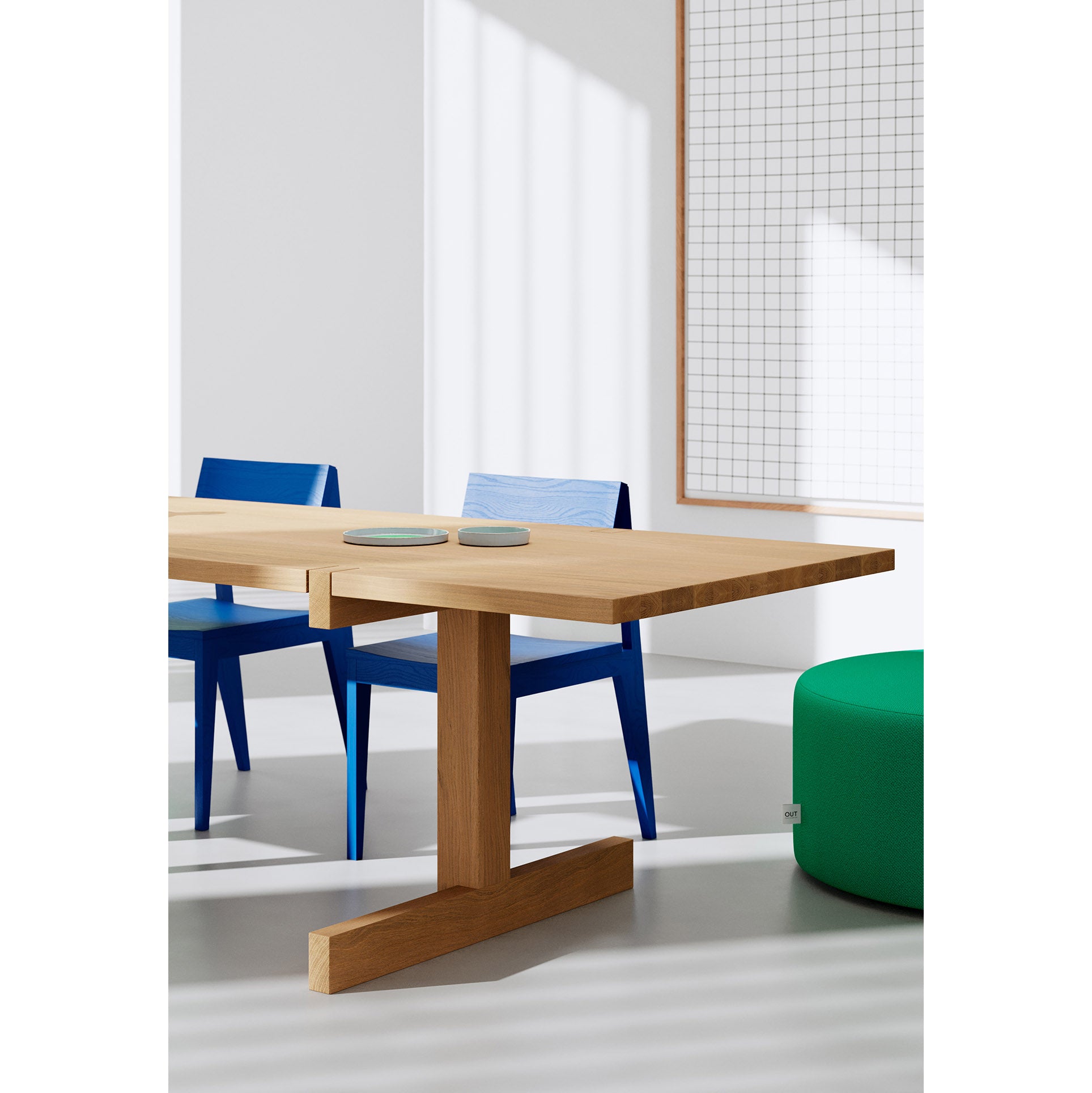 A minimalist dining scene showcases an Objekte unserer Tage Richter Table, crafted from sustainable forestry, with two blue chairs. Nearby, a green ottoman rests. Sunlight streams through a window onto large grid-patterned artwork on the wall, casting gentle shadows.