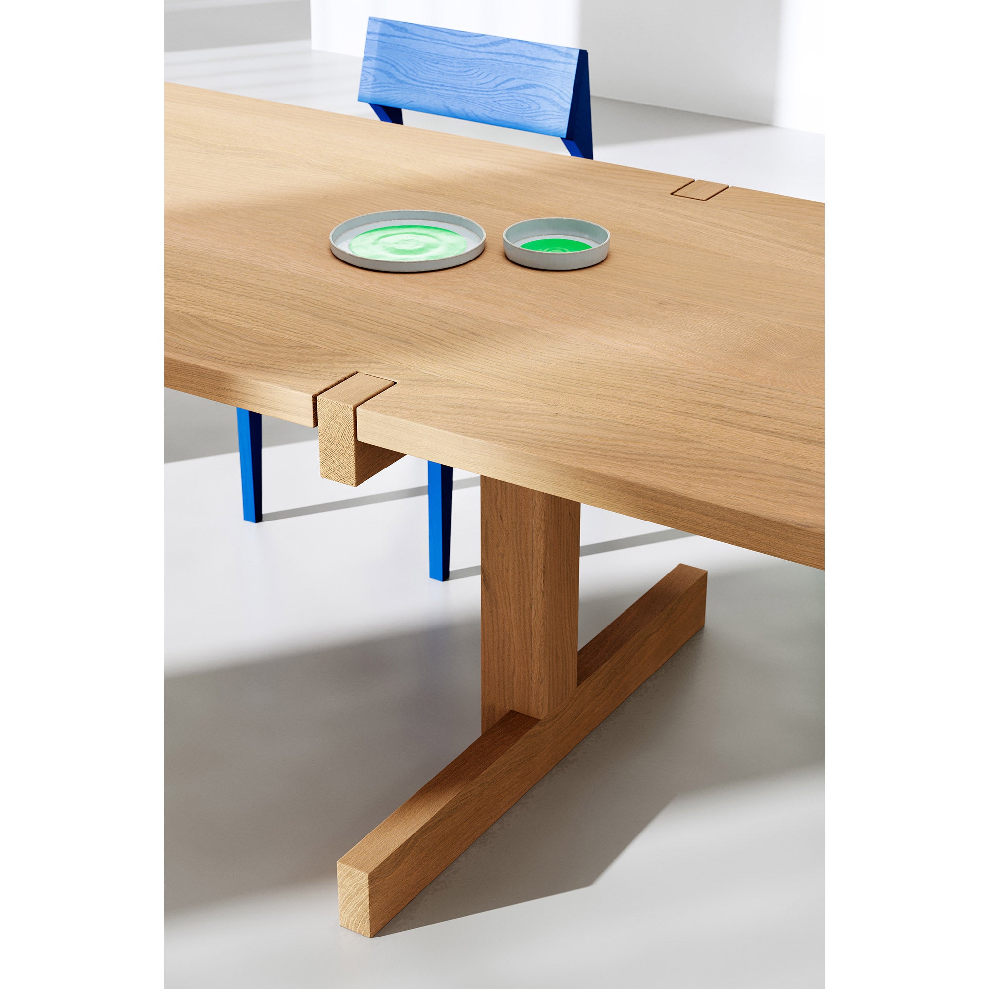 The Richter Table by Objekte unserer Tage features a modern, minimalist design with unique joints. Complemented by a bright blue chair and two small green dishes, this sustainable forestry-crafted setting highlights the furnitures simple elegance.