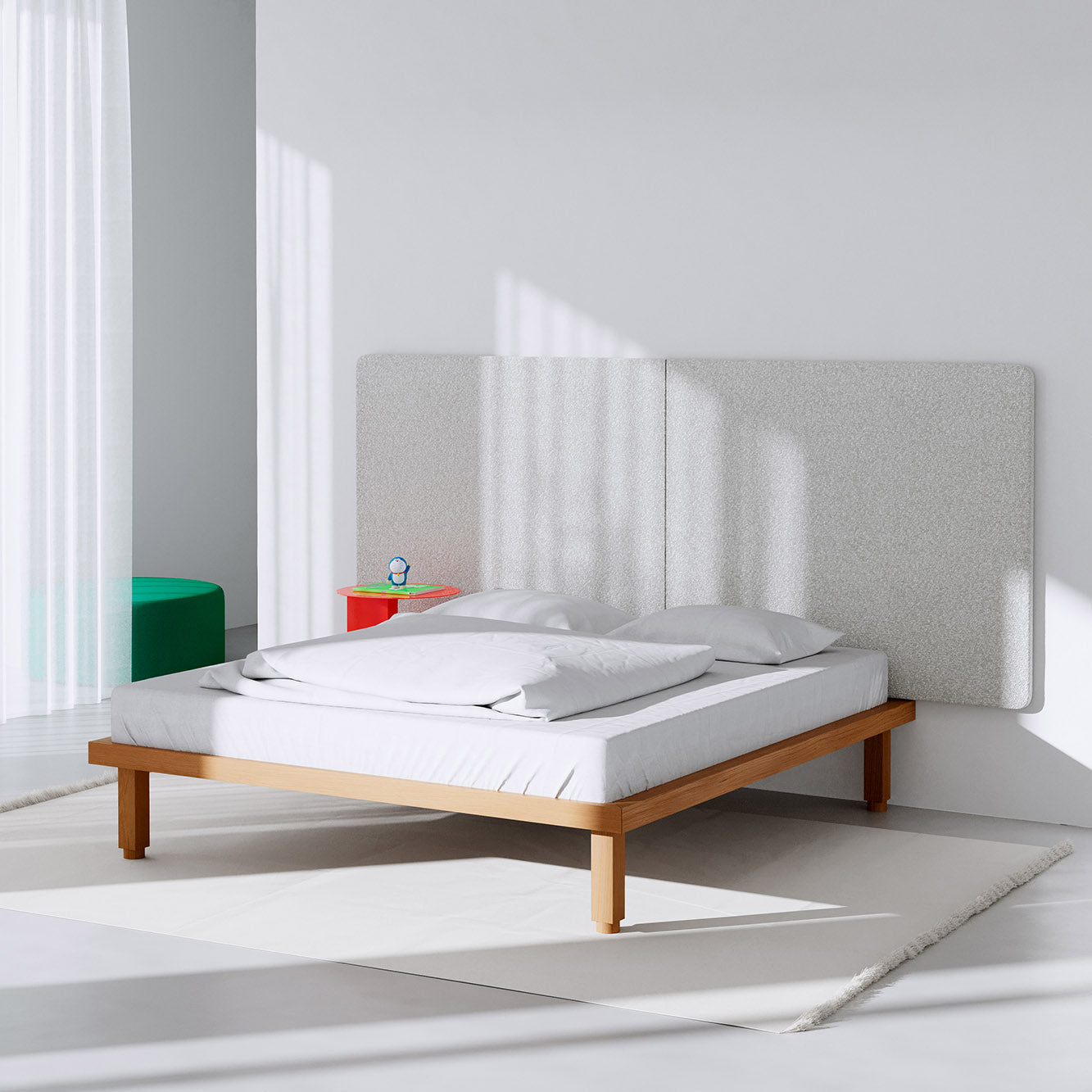A minimalist bedroom showcases the Kaya Bed by Objekte unserer Tage, crafted sustainably. White bedding contrasts with a light-textured wall. Nearby, a small red side table holds a clock, while a green cylindrical stool adds interest. Sunlight filters through sheer curtains, creating an ideal sleep space.