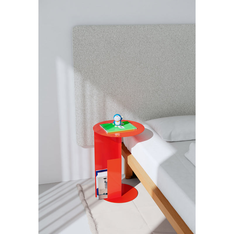 A modern, minimalist bedroom showcases the Objekte unserer Tage KAYA Bed with a white and gray headboard. A red side table holds a toy and a green book, inviting restful sleep. Sunlight casts gentle shadows on the sustainably sourced rug.