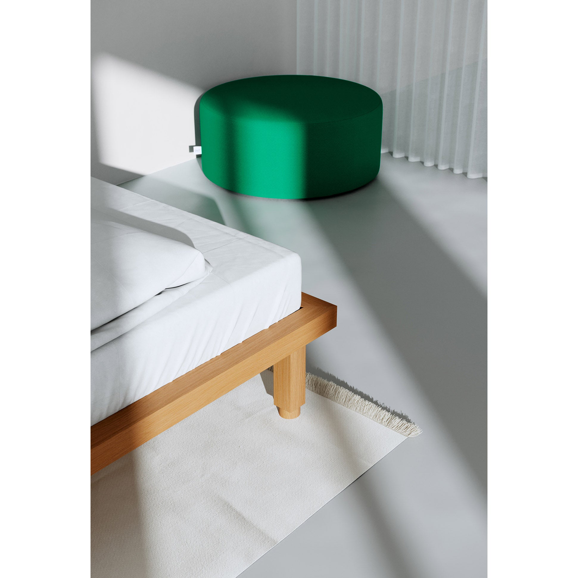 A minimalist room features the Kaya Bed by Objekte unserer Tage, crafted sustainably. White bedding lies to the left, and a textured rug peeks underneath. In the background, a large green ottoman complements white curtains, enhancing tranquility as shadows dance around.