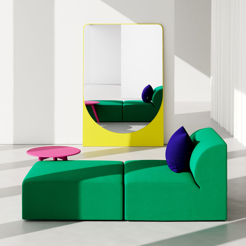 A modern living room showcases a green sectional sofa with purple cushions. Beside it, a round pink coffee table enhances the decor, while a large yellow-framed FRIEDRICH MAX Mirror by Objekte unserer Tage reflects its sculptural allure against white walls and bright lighting.