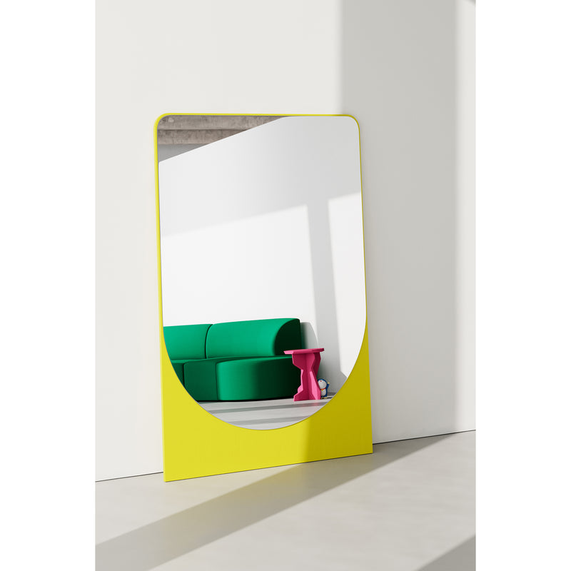 A modern room showcases the FRIEDRICH MAX Mirror by Objekte unserer Tage, featuring a large yellow frame on a white wall. It reflects a green sofa and pink side table, with sunlight casting shadows that enhance the minimalist interiors sculptural character through its harmonious colors and shapes.