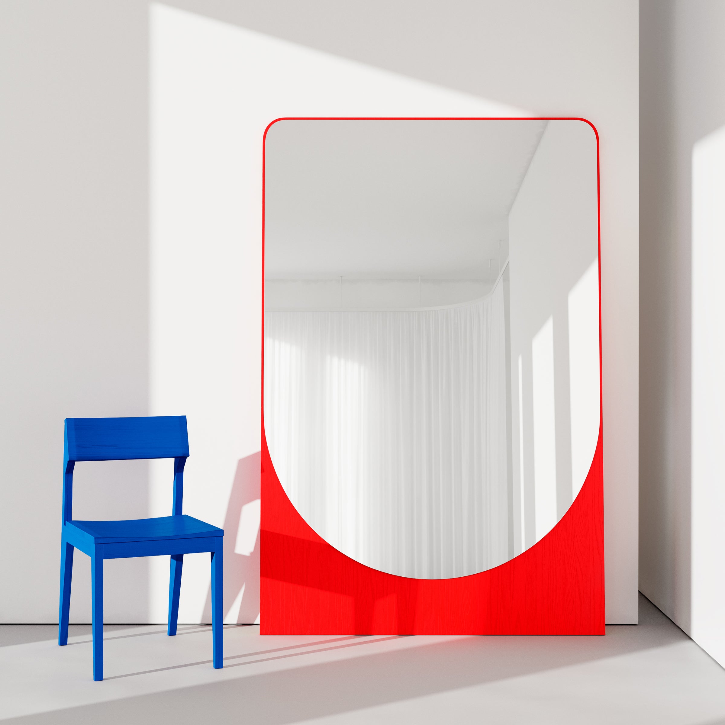A blue chair sits next to the FRIEDRICH MAX Mirror by Objekte unserer Tage, featuring a bold red frame. The mirror leans against a white wall, casting reflections of soft natural light and white curtains in a whimsical style.