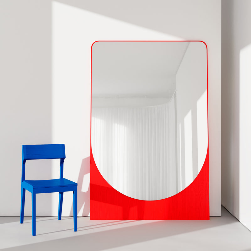 A blue chair sits next to the FRIEDRICH MAX Mirror by Objekte unserer Tage, featuring a bold red frame. The mirror leans against a white wall, casting reflections of soft natural light and white curtains in a whimsical style.