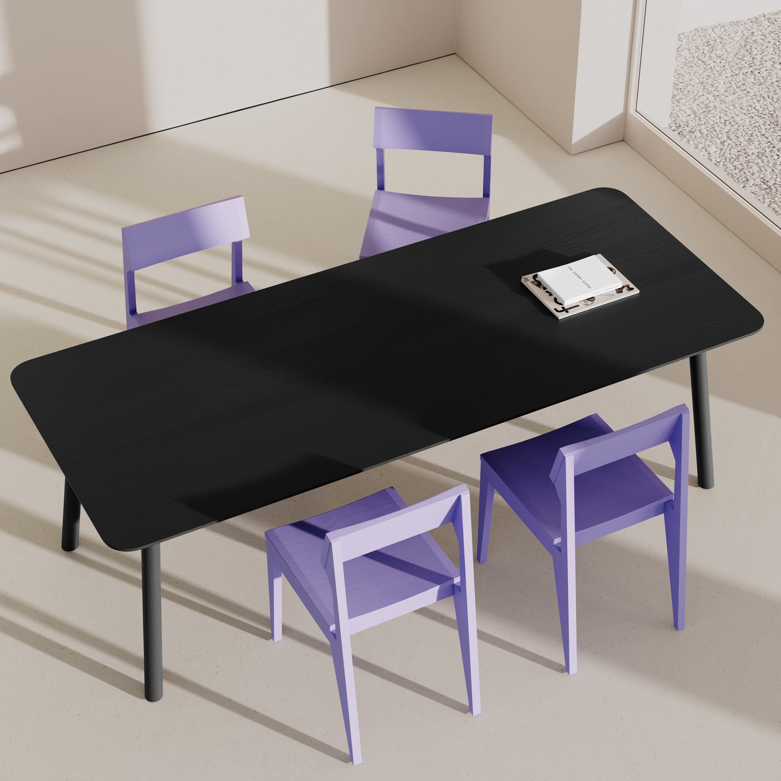 The modern dining room features the Meyer Color Table by Objekte unserer Tage, a rectangular black dining table with an ash wood veneer, surrounded by four purple chairs. Sunlight and large windows highlight its minimalist design and vibrant accents. A closed book sits on the table as shadows play across the light floor.