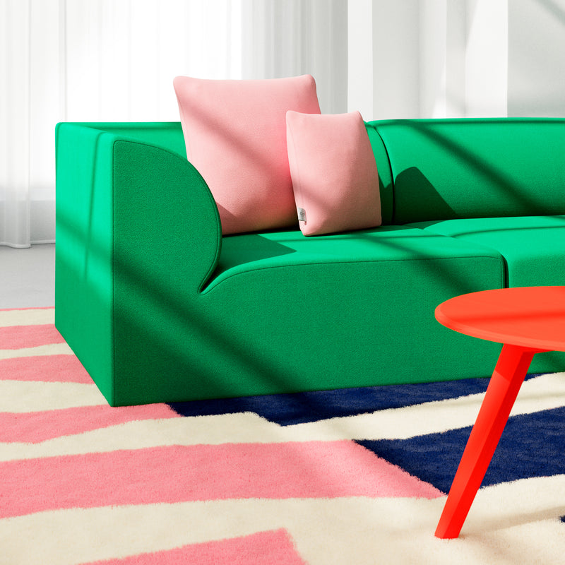 A modern living room showcases an Objekte unserer Tage Weber Modular Sofa in vibrant green with two pink cushions. A carpet displays pink, white, and navy geometric patterns. A small round red table sits before the sofa as sunlight filters through sheer curtains, evoking German sensuality.