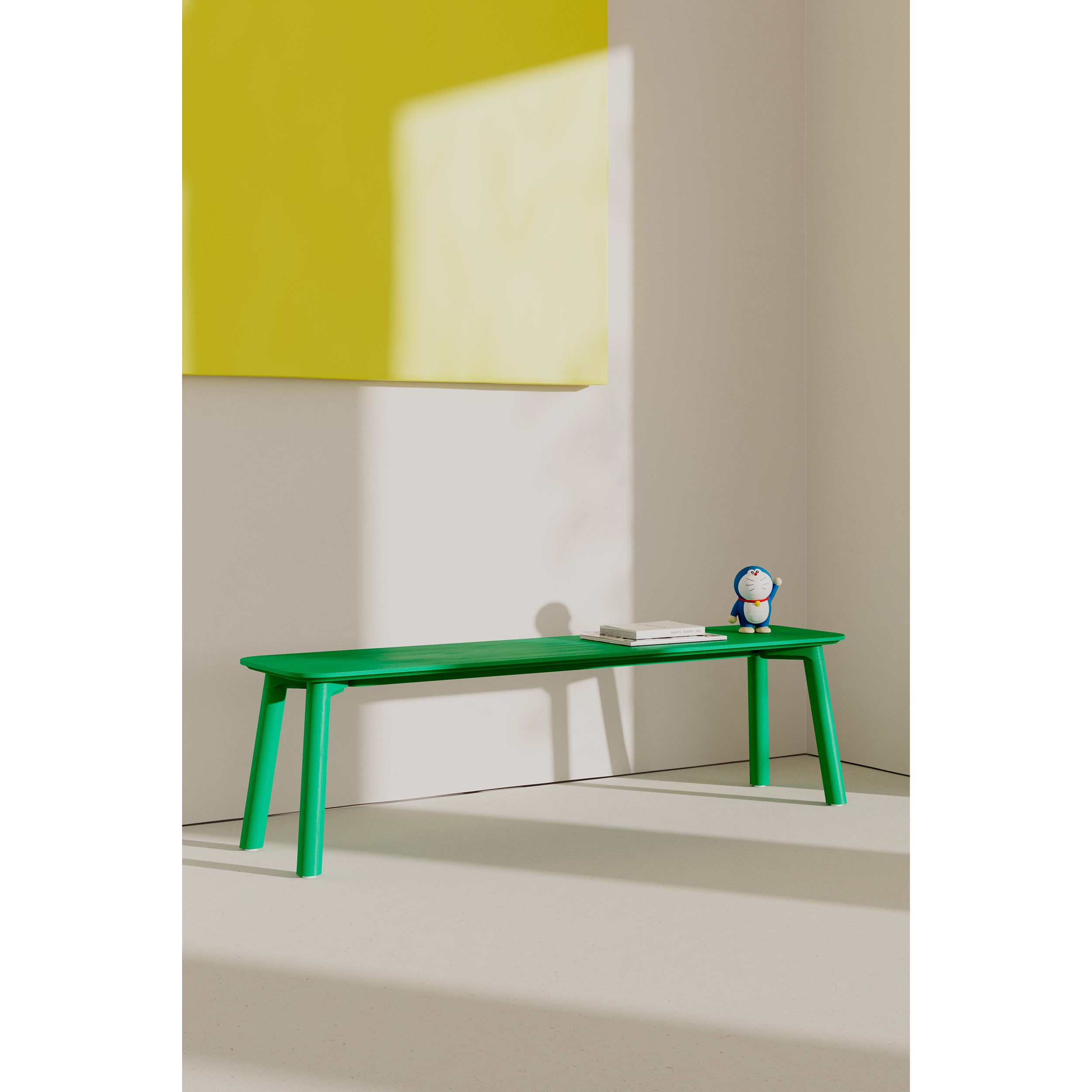 A vibrant green Meyer Color Bench by Objekte unserer Tage rests against a white wall topped with yellow square artwork. An ash wood veneer notebook and a small penguin toy are on the bench, while sunlight filters in, casting shadows across the wall and floor.