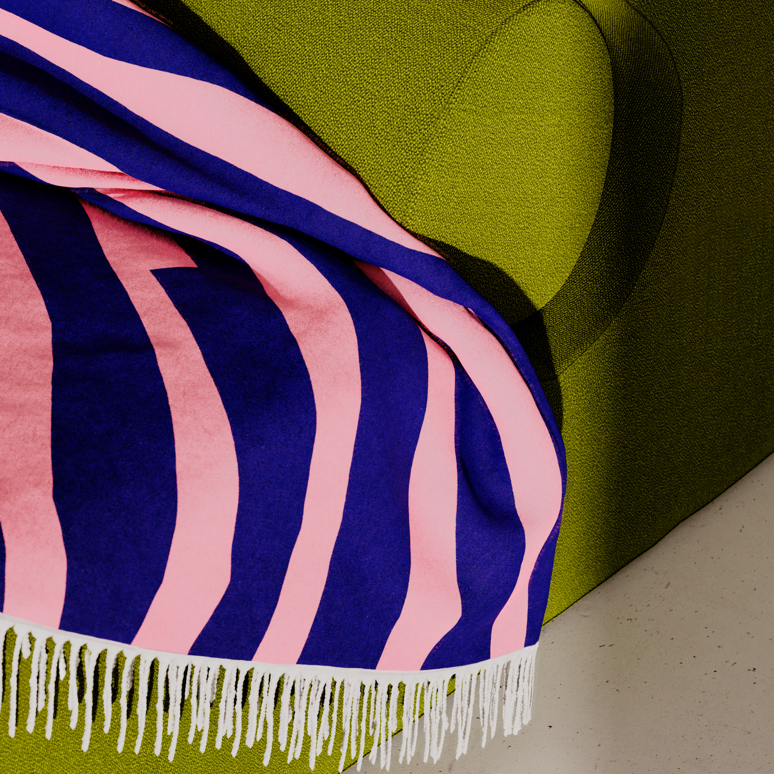 A green couch features a SEIDEL Throw from Objekte unserer Tage, made of organic cotton. The pink and navy striped throw, with fringe edges, adds flair to the textured fabric while snugly resting on the armrest. It beautifully complements the light-colored floor underneath.