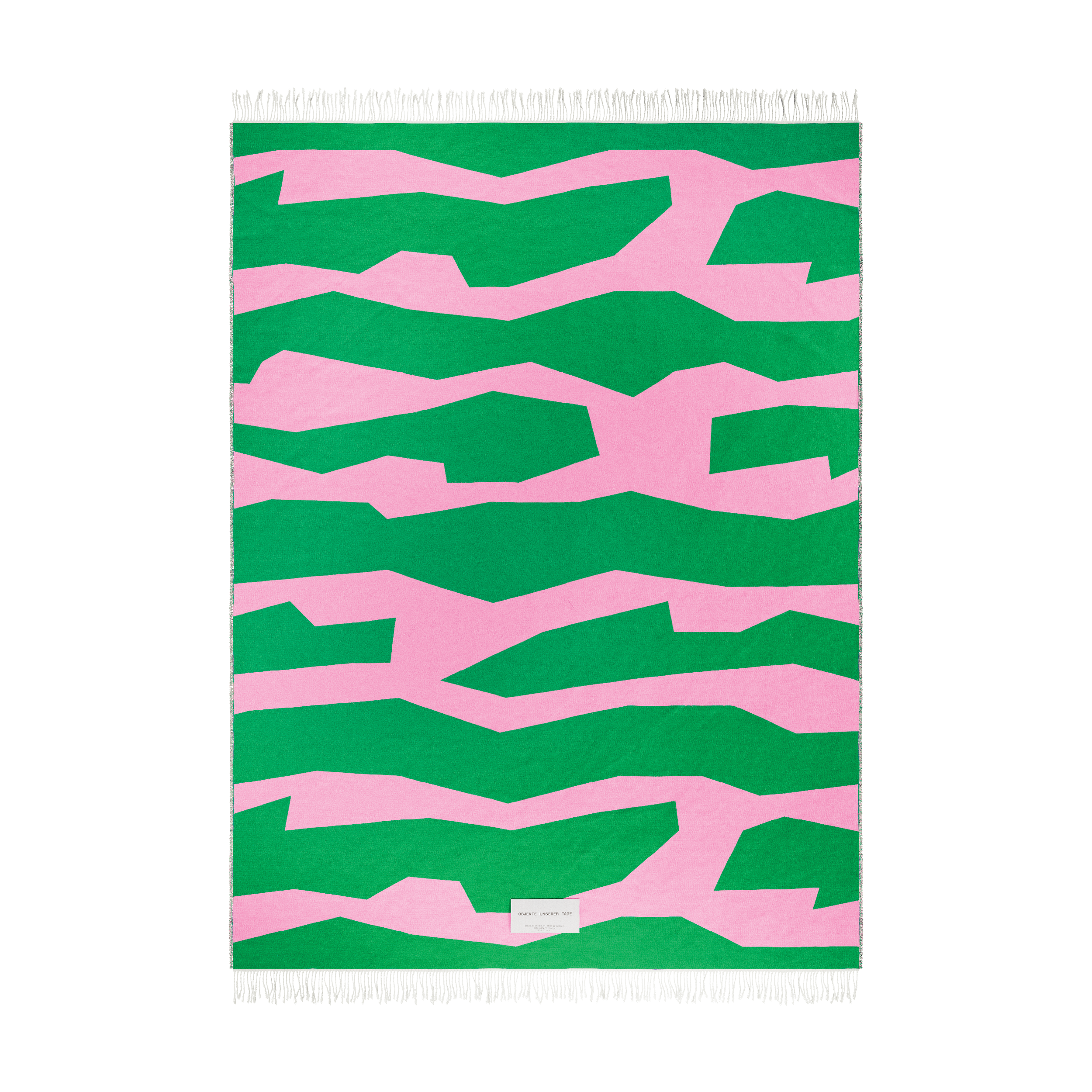 The SEIDEL Throw by Objekte unserer Tage is a rectangular organic cotton rug with an abstract green-and-pink zigzag pattern, featuring bold geometric design on a pink backdrop. It has white fringe on the short ends and a small label near one edge.