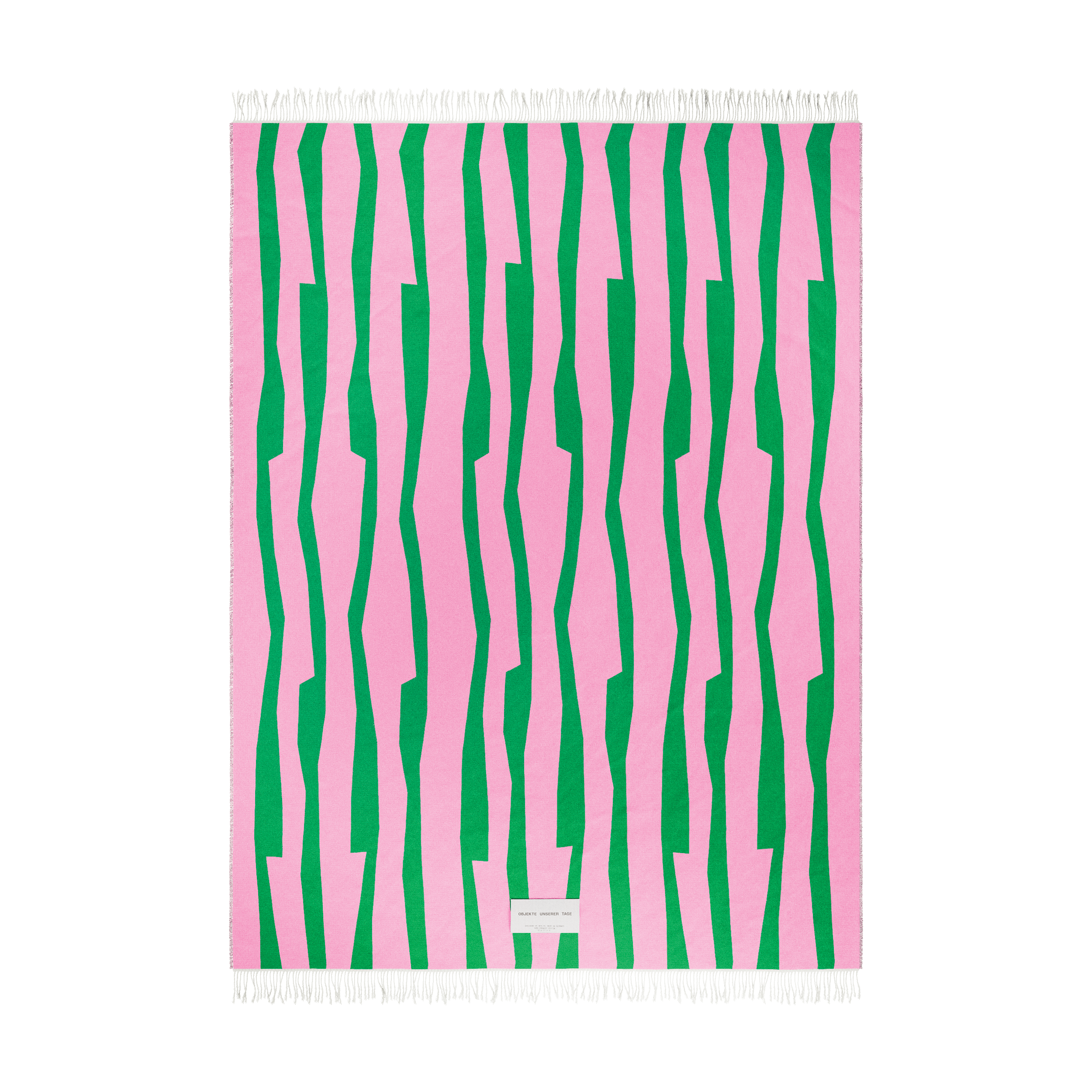 The SEIDEL Throw by Objekte unserer Tage is made from organic cotton, featuring a pink base with vertical green stripes and small white fringes. It has an abstract design with a white patch containing text near the bottom center.