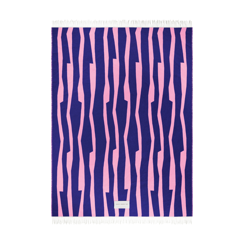 The SEIDEL Throw from Objekte unserer Tage is a rectangular textile made of organic cotton, featuring fringe edges and an abstract graphic design with irregular pink stripes on a dark blue background, along with a small white label at the bottom center.
