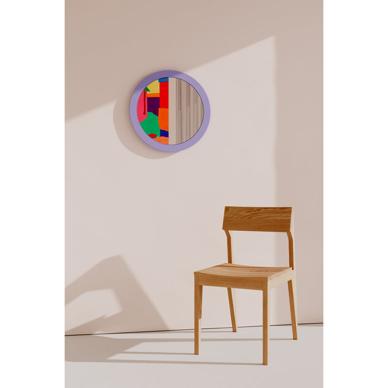 A wooden ash chair stands on a light floor near a beige wall. Above it, the Lorenz Mirror by Objekte unserer Tage with a purple frame reflects a vibrant geometric design. Sunlight adds shadows, enhancing depth and intrigue.