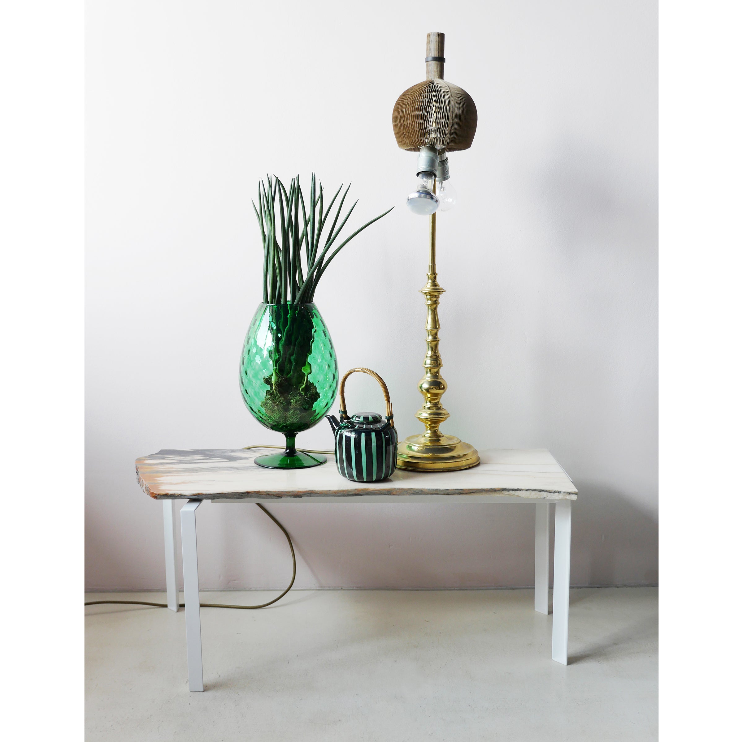 A Petite Table DAngle by Atelier Haussmann, featuring white legs, displays a green glass vase with long leaves, a small striped ceramic pot, and a tall ornate brass lamp with a brown shade; the setup is against a plain white wall.