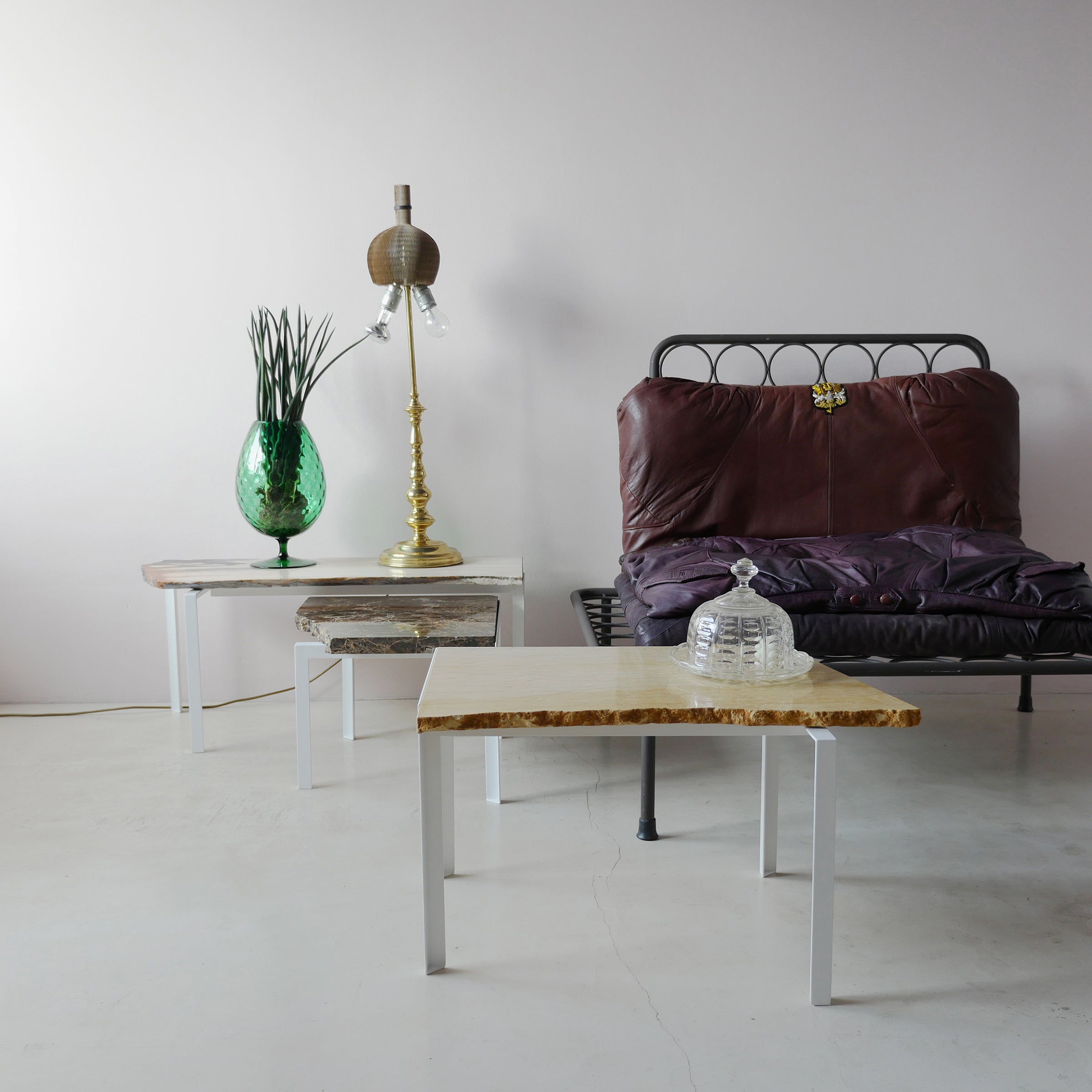 A minimalistic interior features a metal bench with a brown cushion beside Atelier Haussmanns Petite Table DAngle, topped with a green glass vase and unique gold lamp. A nearby table displays a decorative glass object on marble, all set against a light gray wall.