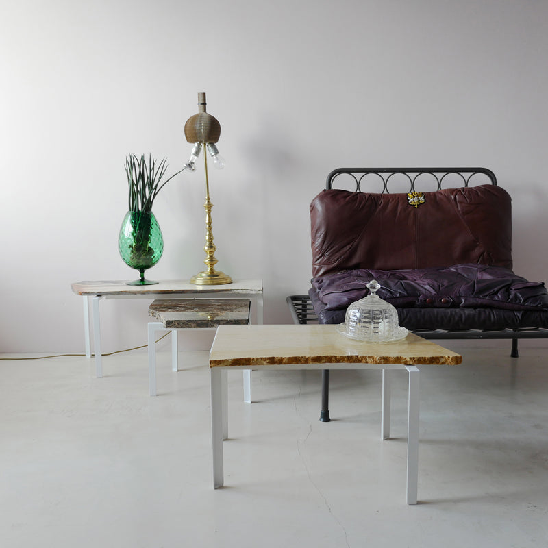 A minimalistic interior features a metal bench with a brown cushion beside Atelier Haussmanns Petite Table DAngle, topped with a green glass vase and unique gold lamp. A nearby table displays a decorative glass object on marble, all set against a light gray wall.