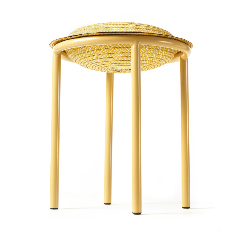 The Ames Cana Stool, in a light yellow tone, showcases a round woven wicker seat with four sturdy legs, photographed from a low angle against a pristine white background. Its unique design makes this stool stand out despite its subtle color.